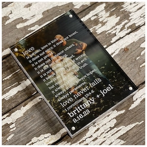 May include: A clear acrylic photo block with a printed image of a couple. The text "love never fails" and the Bible verse 1 Corinthians 13:4-8 are printed in white. The names "brittany + joel" and the date "9.16.23" are also printed.