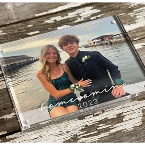 May include: A clear acrylic photo frame with a picture of a couple in formal attire. The photo is taken outdoors with a body of water in the background. The text "homecoming 2023" is written in white cursive on the bottom of the frame.