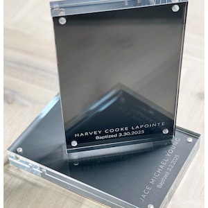 May include: Clear acrylic photo frame with a black insert. The frame is held together with small silver screws. The text "HARVEY COOKE LAPOINTE Baptized 3.30.2025" and "JACE MICHAEL YOUNG Baptized 3.2.2025" are visible.