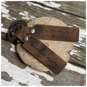 May include: Two brown leather keychains with metal rings. Each keychain has a small metal rivet and a pattern of dots and dashes, resembling Morse code.