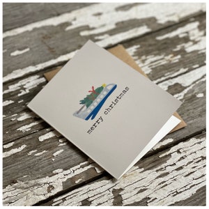 May include: A gray card with a blue and white airplane carrying a green Christmas tree with red ribbon and a yellow star on top. The card says "merry christmas" in black text.