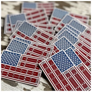 May include: A collection of rectangular stickers featuring a stylized American flag design. The flag's blue field is filled with white airplane silhouettes, and the red stripes depict airport runways. The stickers are arranged on a distressed, white wooden surface.