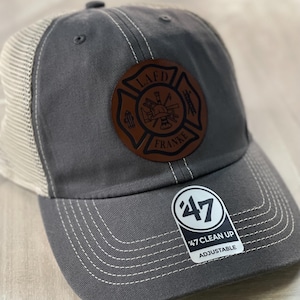 May include: Gray and beige trucker hat with a brown leather patch featuring a fire department logo with the text "LAFD FRANKE". The hat has a white "47 CLEAN UP ADJUSTABLE" label.