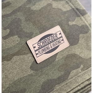 May include: A brown leather patch with white stitching and black text that reads "Schroeder Plumbing & Heating" on a green camouflage fabric background.