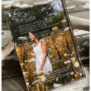 May include: A clear acrylic photo frame with a white background and a poem printed on it. The poem is about letting go and moving on. The photo in the frame is of a person standing in a field of yellow flowers.