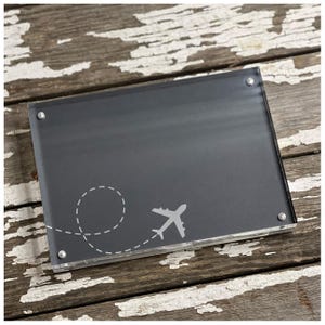 May include: A black acrylic photo frame with a white airplane silhouette and a dashed line representing a flight path. The frame is on a wooden surface.