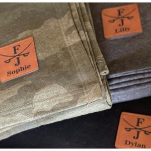 May include: Close-up of folded blankets in shades of green camouflage and gray. Each blanket has a square, brown leather patch with the initials "FJ", crossed swords, and a name: Sophie, Lilly, or Dylan.