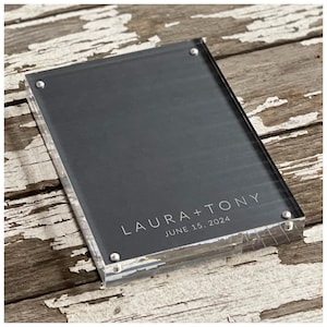 May include: A clear acrylic photo frame with a black background and silver accents. The frame has the text "LAURA + TONY" and "JUNE 15, 2024" printed on it.