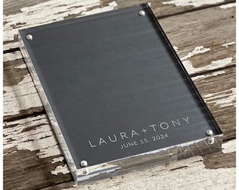 May include: A clear acrylic photo frame with a black background and silver accents. The frame has the text "LAURA + TONY" and "JUNE 15, 2024" printed on it.