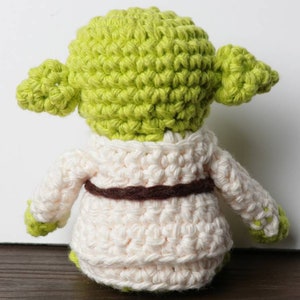Pu&ograve; includere: Una bambola di Yoda all'uncinetto fatta a mano, vista da dietro. La bambola presenta una testa verde chiaro con grandi orecchie, una veste color crema e una cintura marrone. Le mani e i piedi sono verdi. La bambola &egrave; realizzata con un punto all'uncinetto stretto.