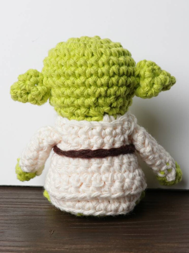 Pu&ograve; includere: Una bambola di Yoda all'uncinetto fatta a mano, vista da dietro. La bambola presenta una testa verde chiaro con grandi orecchie, una veste color crema e una cintura marrone. Le mani e i piedi sono verdi. La bambola &egrave; realizzata con un punto all'uncinetto stretto.
