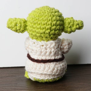Pu&ograve; includere: Bambola di Yoda all'uncinetto fatta a mano, vista da dietro. La bambola presenta una testa e orecchie verde chiaro, una veste color crema e una cintura marrone. La base della bambola &egrave; verde. La bambola &egrave; su una superficie marrone.
