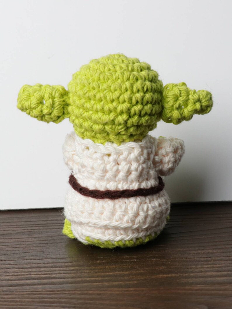 Pu&ograve; includere: Bambola di Yoda all'uncinetto fatta a mano, vista da dietro. La bambola presenta una testa e orecchie verde chiaro, una veste color crema e una cintura marrone. La base della bambola &egrave; verde. La bambola &egrave; su una superficie marrone.
