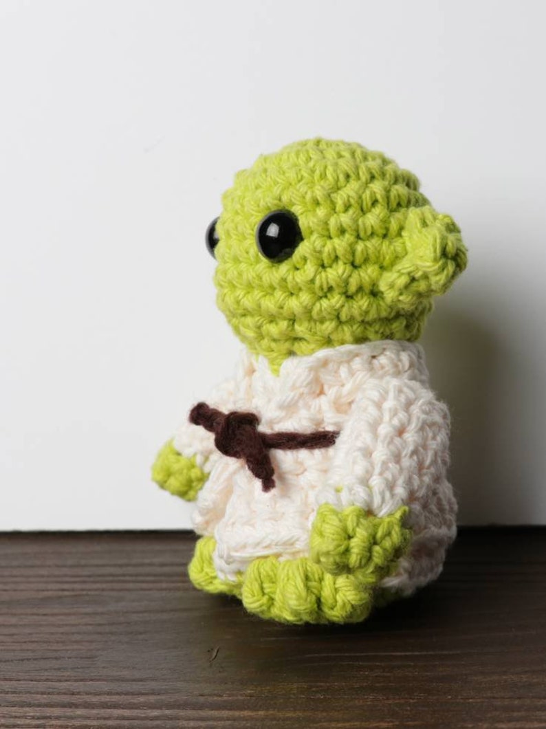 Pu&ograve; includere: Figura all'uncinetto di un personaggio con grandi occhi neri, pelle verde e orecchie a punta. Indossa una veste bianca con una cintura marrone. L'oggetto &egrave; su una superficie di legno.