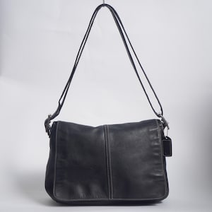 May include: A black leather shoulder bag featuring a fold-over flap, a central vertical seam with white contrast stitching, and a long adjustable strap. A small black leather rectangular tag hangs from a silver-toned metal chain on the side.