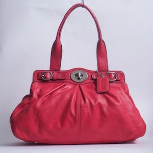 May include: A bright red leather handbag featuring a central silver-toned turn-lock closure, two top handles, and decorative side buckles. A small rectangular leather tag hangs from the right side of the bag, which has a gathered, pleated design.