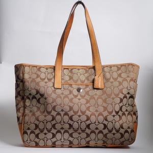 May include: A tan and brown canvas tote bag featuring a repeating C logo pattern, tan leather trim, two long shoulder straps, a silver-toned metal snap closure, and a small leather luggage tag hanging from one of the straps.