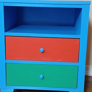 May include: A blue wooden nightstand with three drawers. The top drawer is red, the middle drawer is green, and the bottom drawer is blue. The nightstand has a shelf above the drawers.
