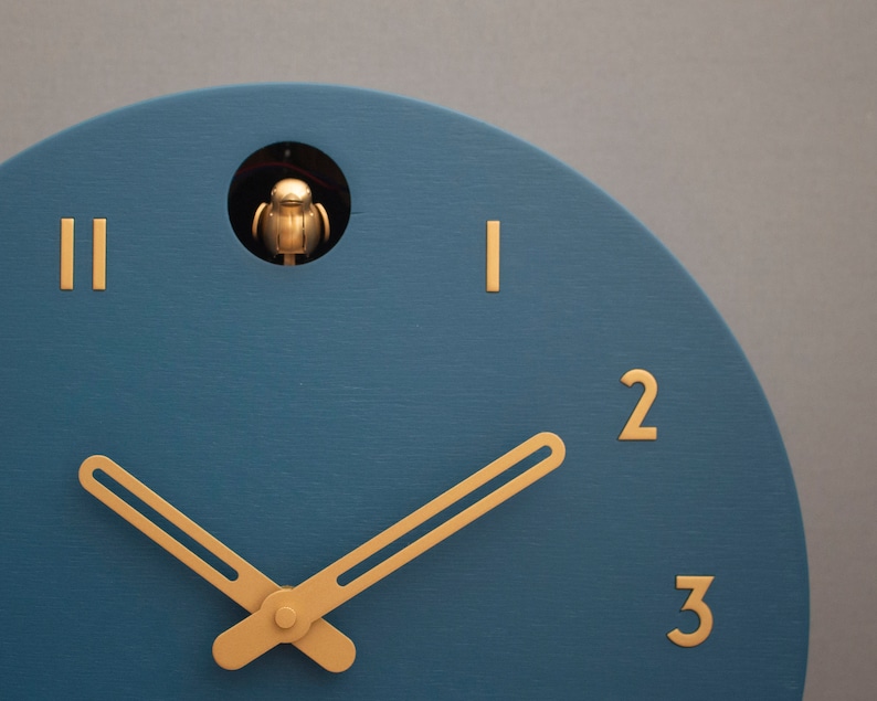 May include: A blue cuckoo clock with gold hands and Roman numeral markings. The clock face has a small opening at the top with a gold bird figurine inside.