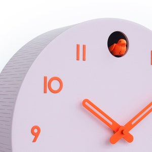 May include: A modern, pastel pink cuckoo clock with orange accents. The clock features orange numerals, hands, and a small bird inside a circular opening. The clock's face is a soft pink, and the clock has a textured edge.