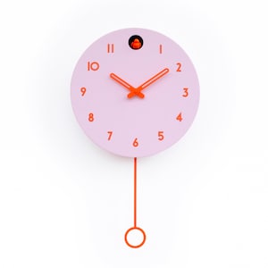 May include: A round, pink cuckoo clock with orange hands, numbers, and pendulum. The clock has a small orange bird inside a circular opening at the top. The pendulum hangs from the bottom of the clock.