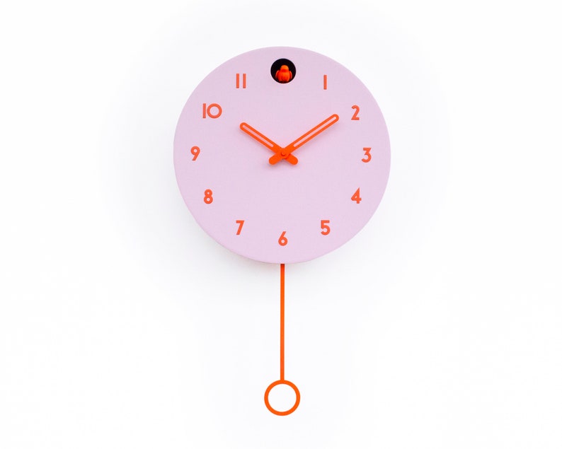 May include: A round, pink cuckoo clock with orange hands, numbers, and pendulum. The clock has a small orange bird inside a circular opening at the top. The pendulum hangs from the bottom of the clock.