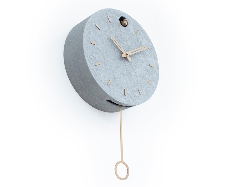 May include: A grey round clock with a pendulum and gold-coloured hands. The clock has a textured surface and a small hole at the top.