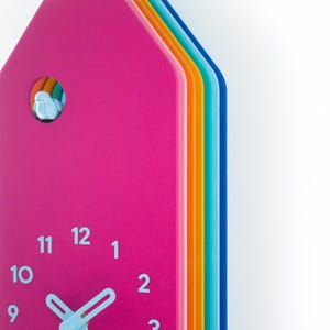 Könnte beinhalten: Eine farbenfrohe Kuckucksuhr mit einer rosafarbenen Vorderseite und einem Regenbogen an Farben an der Seite. Das Zifferblatt der Uhr hat weiße Zahlen und Zeiger.