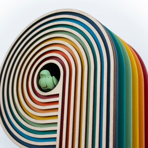 May include: A wooden rainbow arch with multiple layers of colorful wood, each layer a different color. A small green bird figurine is nestled in the opening of the arch.
