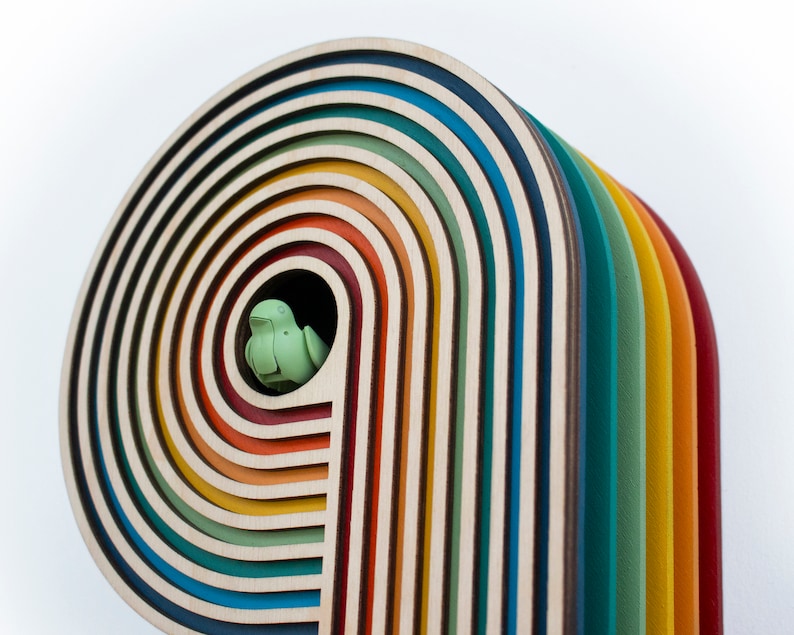 May include: A wooden rainbow arch with multiple layers of colorful wood, each layer a different color. A small green bird figurine is nestled in the opening of the arch.