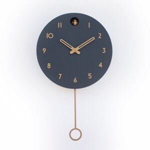 May include: A dark blue cuckoo clock with gold-coloured hands and numbers. The clock has a pendulum and is hanging on a white wall.