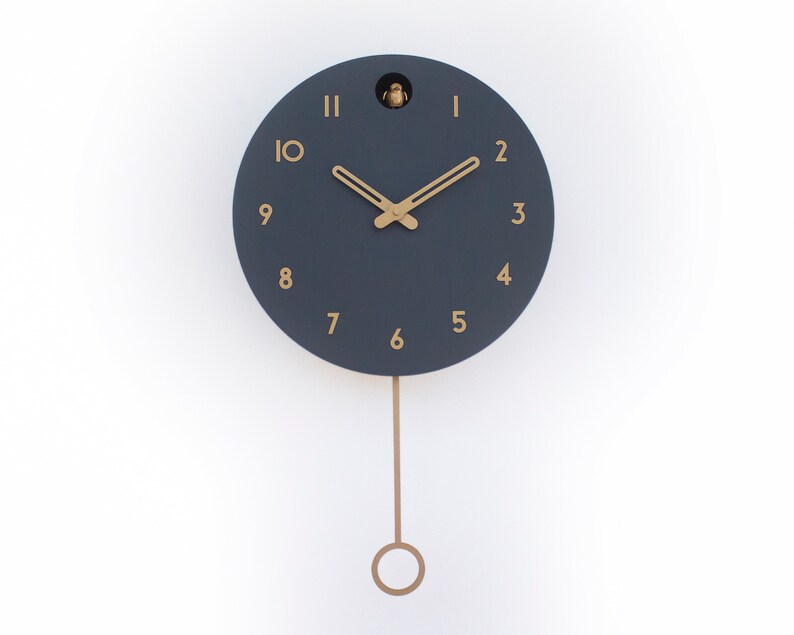 May include: A dark blue cuckoo clock with gold-coloured hands and numbers. The clock has a pendulum and is hanging on a white wall.