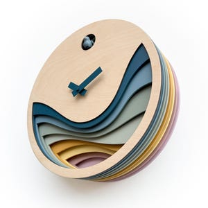 K&ouml;nnte beinhalten: Eine runde Wanduhr aus Holz mit einem geschichteten Design, das blaue, gr&uuml;ne, gelbe und rosa Wellen zeigt. Die Uhr hat einen Kuckuck und ein einfaches schwarzes und blaues Zifferblatt.