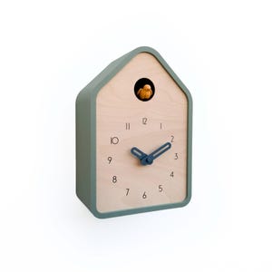 May include: A modern cuckoo clock in a house shape. The clock has a light wood face with black numbers and blue hands. The frame is a muted green color. A small yellow bird is visible in the cuckoo's opening.
