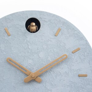 May include: A light blue round clock face with gold-coloured hour and minute hands. The clock has a small opening at the top with a gold-coloured bird figurine inside.