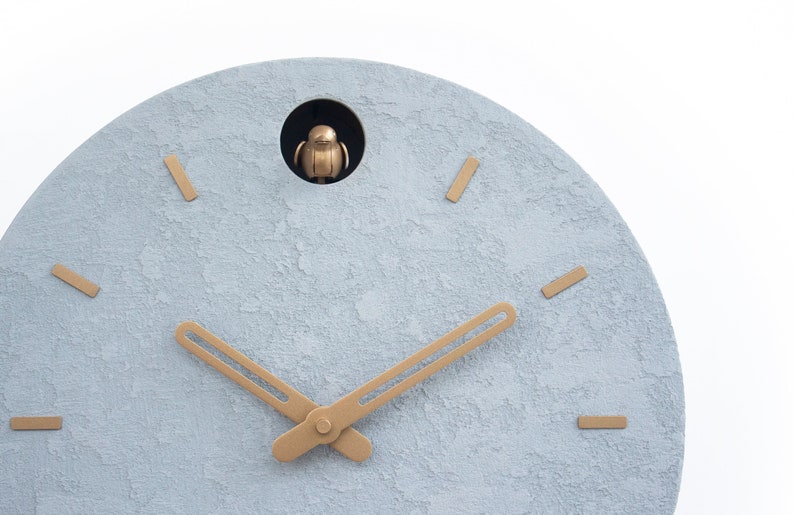 May include: A light blue round clock face with gold-coloured hour and minute hands. The clock has a small opening at the top with a gold-coloured bird figurine inside.