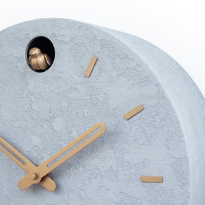 May include: A grey concrete cuckoo clock with gold-coloured hands and a small bird that pops out of the hole at the top of the clock.