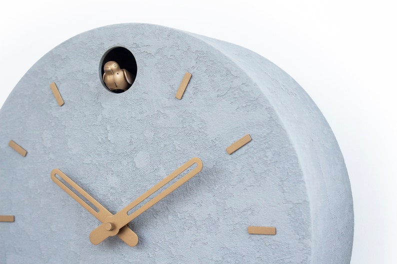 May include: A grey concrete cuckoo clock with gold-coloured hands and a small bird that pops out of the hole at the top of the clock.