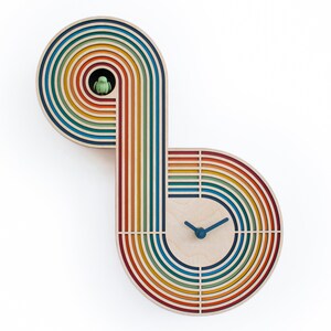 May include: A rainbow-colored wooden clock with a unique design. The clock face is in the center of the design, and the hands are blue. The clock is made of wood and has a rainbow-colored design.