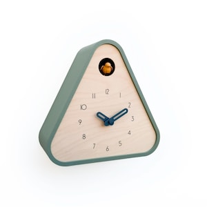 May include: A triangular cuckoo clock with a light wood face and a sage green frame. The clock features black numerals, blue hands, and a small yellow bird. The clock is a modern take on a classic design.
