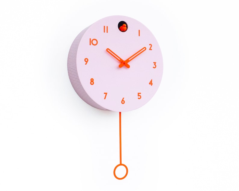 May include: A round, light pink wall clock with orange numerals and hands. The clock has a small orange bird inside a hole at the top and an orange pendulum hanging below. The clock is mounted on a white wall.