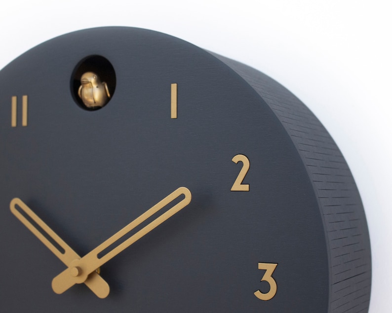 May include: A dark grey cuckoo clock with gold-coloured hands and Roman numeral markings. The clock has a small bird that pops out of the top.