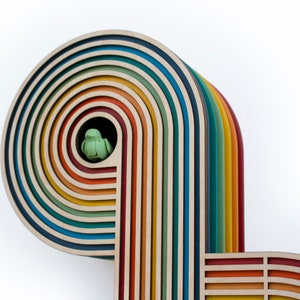 May include: A wooden rainbow arch with multiple layers of colorful wood. The arch is made of different shades of brown, orange, yellow, green, blue, and purple. A small green bird figurine is perched inside the arch.