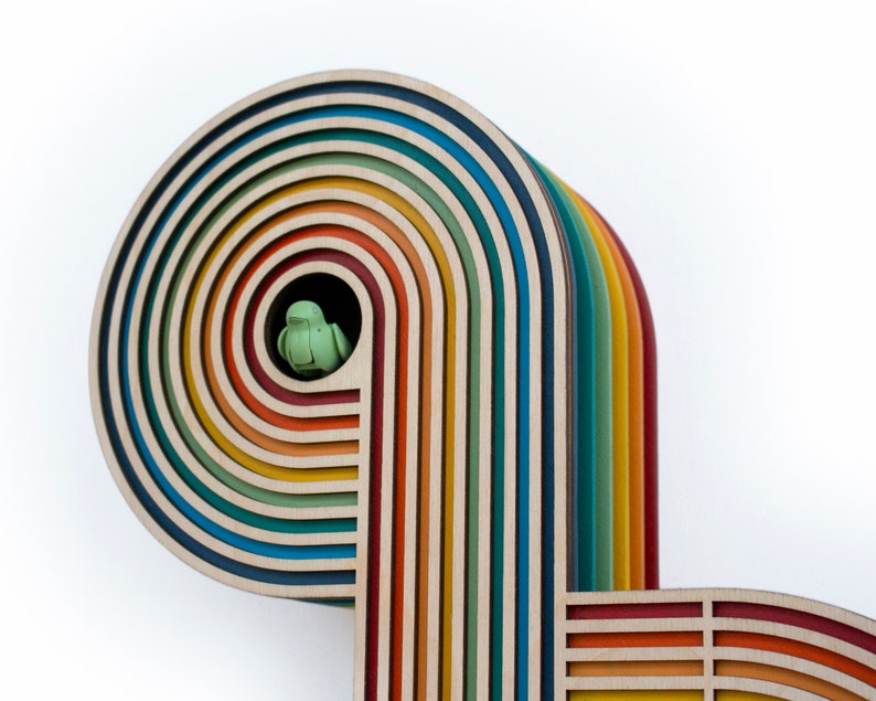 May include: A wooden rainbow arch with multiple layers of colorful wood. The arch is made of different shades of brown, orange, yellow, green, blue, and purple. A small green bird figurine is perched inside the arch.