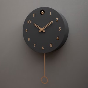 May include: A black cuckoo clock with gold-coloured hands and numbers. The clock has a pendulum and is hanging on a grey wall.