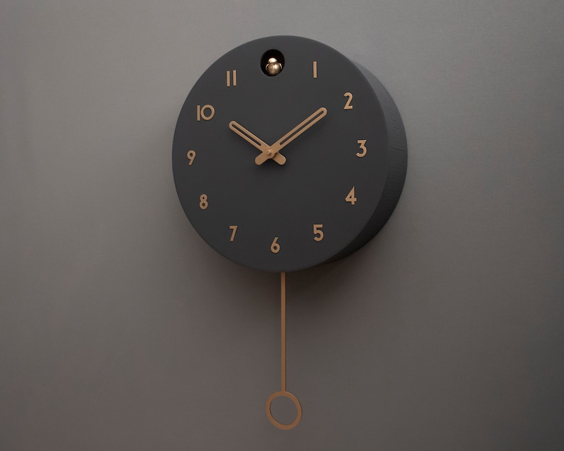 May include: A black cuckoo clock with gold-coloured hands and numbers. The clock has a pendulum and is hanging on a grey wall.