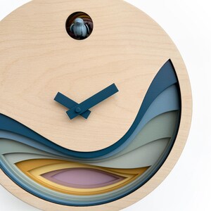 K&ouml;nnte beinhalten: Eine Holzuhr mit einem mehrschichtigen Design in Blau, Gr&uuml;n und Gelb. Das Zifferblatt der Uhr ist wei&szlig; mit schwarzen Zeigern und einem kleinen Vogelausschnitt oben.
