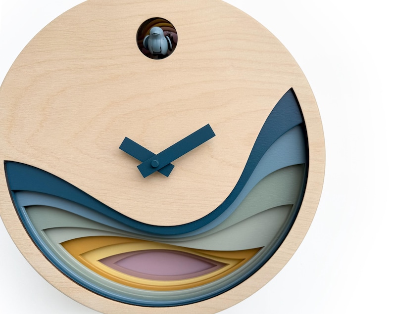 K&ouml;nnte beinhalten: Eine Holzuhr mit einem mehrschichtigen Design in Blau, Gr&uuml;n und Gelb. Das Zifferblatt der Uhr ist wei&szlig; mit schwarzen Zeigern und einem kleinen Vogelausschnitt oben.