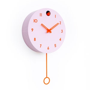 May include: A round, light pink wall clock with orange hands and numerals. The clock has a matching orange pendulum and a small orange bird detail at the top. The clock is mounted on a white wall.
