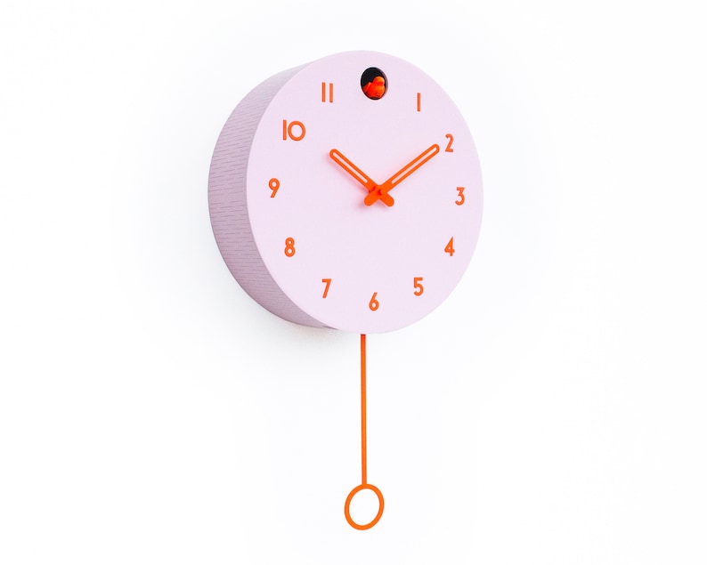 May include: A round, light pink wall clock with orange hands and numerals. The clock has a matching orange pendulum and a small orange bird detail at the top. The clock is mounted on a white wall.